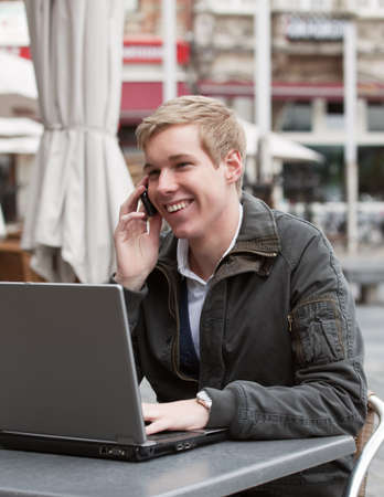 Happy young man speaking on cellphone in a street cafe while working with his laptopの写真素材