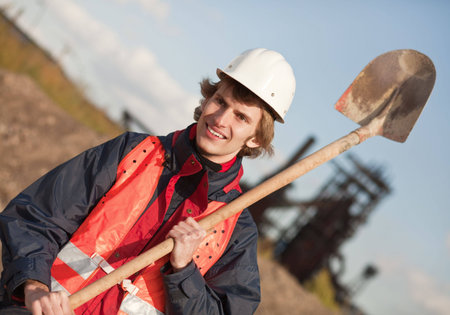 Young manual worker with shovel standing against industrial background