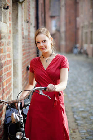 Young blond woman in red dress standing on a street with old bicycleの写真素材