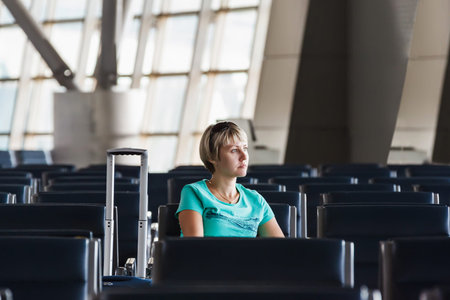 beautiful young woman with blond short hair with a suitcase sitting on a chair at the airport and waiting for her flightの写真素材