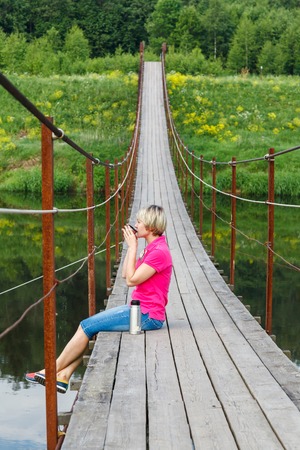 blonde girl with short haircut drinking tea from a thermos on Pedestrian suspension bridge of steel and wood over the river, summer in Russiaの写真素材