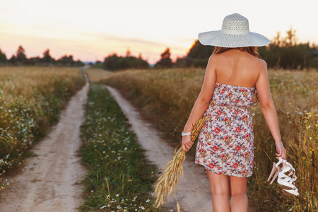 Portrait of young pretty girl at summer weather dressed in bright hat goes on road through a field of wheatの写真素材
