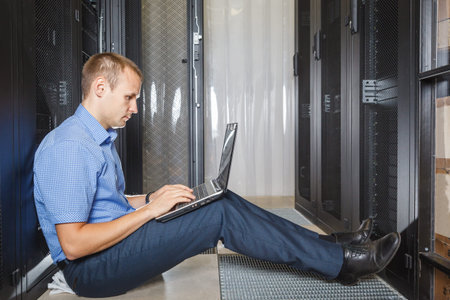 IT Engineer sitting on the floor between the rows in the Data Center and working on laptop