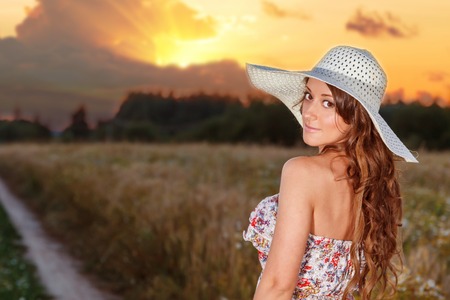 Portrait of young pretty girl at summer weather dressed in bright hat goes on road through a field of wheatの写真素材