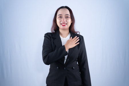 A confident young woman in a black suit places her hand on her chest while smiling, conveying...