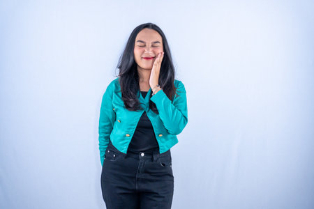 A young woman with closed eyes and a bright smile gently touches her cheek, expressing joy or a playful moment.の写真素材