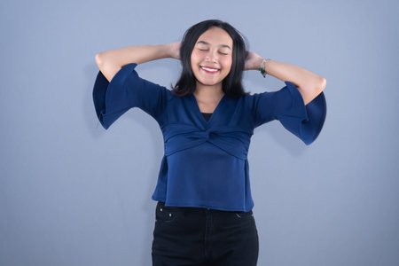A happy Asian woman in a blue shirt smiling with hands behind her head.