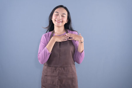A smiling woman with closed eyes and hands on her chest, wearing an apron, against a plain...