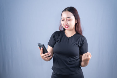 A smiling young woman in a black t-shirt happily holds a smartphone, clenching her fist in...