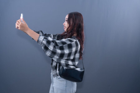 A young woman with long, dark hair takes a selfie with her phone, wearing a plaid shirt and...