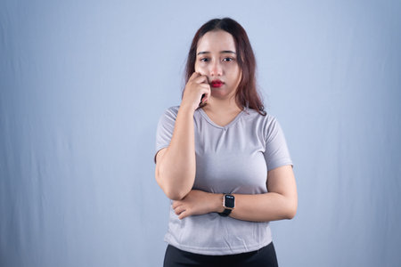 Young Asian woman in grey t-shirt, looking sad or thoughtful, hand near face, on grey background