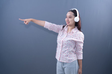 Young Asian woman with headphones, smiling and pointing left, on a grey background