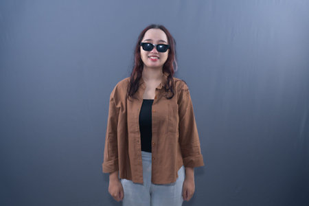 A young Asian woman in a brown shirt, black top, and sunglasses, smiling, on a grey background