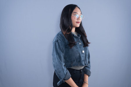Young Asian woman in denim jacket and glasses, looking right with a gentle smile, on grey background