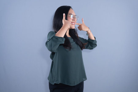 Young Asian woman in green top & glasses, pointing to her smiling teeth, on grey background