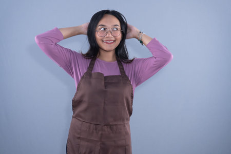 A young Asian woman in a purple shirt and brown apron, smiling with hands behind her head