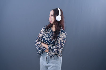 Young Asian woman in floral top, headphones, smiling, arms crossed, looking right, grey background