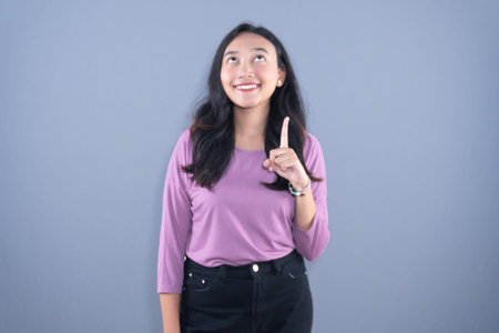 A smiling young woman in a purple shirt and black pants pointing one finger up.