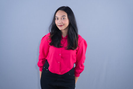 A young woman in a bright pink shirt and black pants smiling faintly in front of a gray background.の写真素材