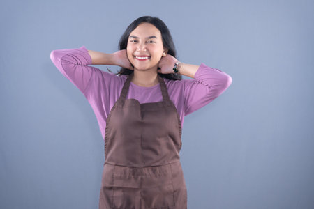 A smiling young woman in a purple shirt and brown apron with her hands behind her head.の写真素材