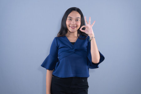 A smiling young woman in a blue shirt is making an OK gesture with her left hand.の写真素材