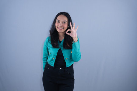 A young woman in a teal jacket smiling and making an OK gesture with her hand.の写真素材