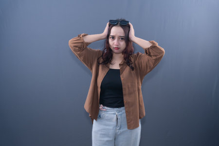 A woman with sunglasses on her head, wearing a brown shirt and jeans, holds her head with both hands.の写真素材