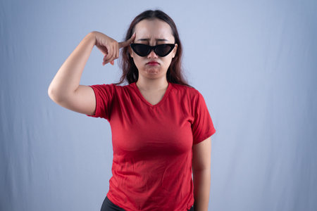 A woman in sunglasses and a red shirt points to her temple with a pouting expression.の写真素材