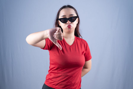 A woman in a red shirt and sunglasses gives a thumbs-down gesture with a pouting expression.