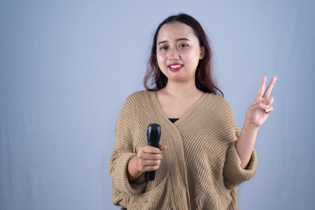 A smiling woman in a beige cardigan holds a microphone and makes a peace sign.の写真素材