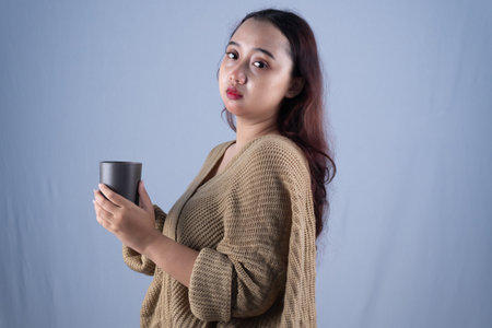 A woman with a serious expression, wearing a brown cardigan, holds a dark gray mug.の写真素材