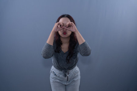 A woman in a gray shirt and light-colored jeans makes a binoculars gesture with her hands over her eyes.の写真素材