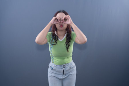 A woman in a green shirt and jeans makes a binoculars gesture with her hands over her eyes.の写真素材