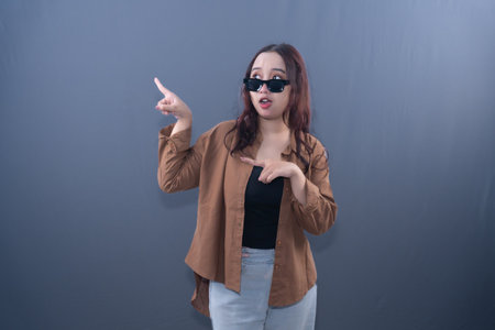 A woman with sunglasses points with both hands, looking surprised. She wears a brown shirt and black top.の写真素材