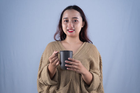 A smiling woman in a brown knitted sweater holds a dark mug with both hands.の写真素材