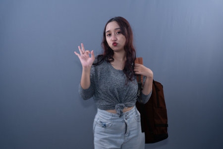 A woman in a gray top and jeans makes an OK sign, holding a brown backpack.の写真素材