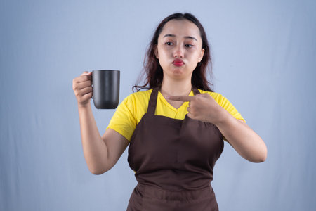 A woman in a yellow shirt and apron points to a dark mug with a pouting expression.の写真素材