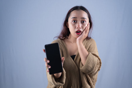 A shocked woman in a beige sweater is holding a smartphoneの写真素材