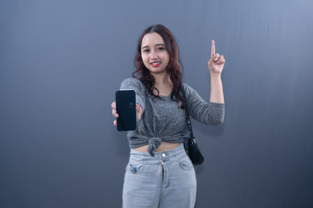 A smiling woman in a gray shirt holds a phone and points upの写真素材
