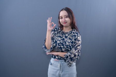 A smiling woman in a navy blue floral top makes an 'okay' hand gesture against a gray backgroundの写真素材