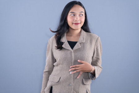 A young woman in a beige blazer looks sideways and smiles while holding her stomachの写真素材