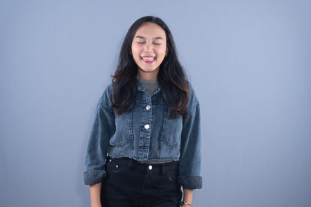 A young woman in a denim jacket smiles with eyes closed, sticking her tongue out playfully