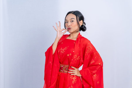 A woman in a red Chinese dress makes an OK sign with a pout against a simple white backgroundの写真素材