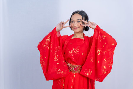 A woman in a red Chinese dress makes double peace signs by her face on a white studio backgroundの写真素材