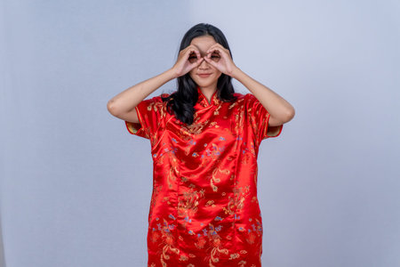 Asian woman in red cheongsam makes okay sign over eyes, looking like glassesの写真素材