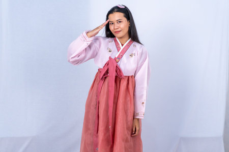 A woman in pink and red Korean Hanbok salutes with a smile against a simple white studio backgroundの写真素材