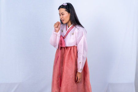 A woman in pink and red Korean Hanbok coughs into her fist against a simple white studio backgroundの写真素材