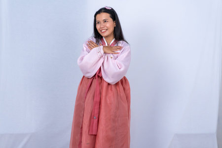 A woman in pink and red Korean Hanbok smiles with arms crossed over her chest on a white backgroundの写真素材