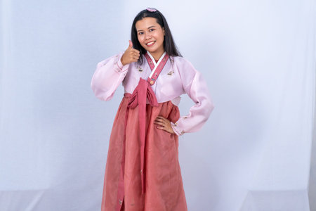 A woman in pink and red Korean Hanbok gives a thumbs up with a smile against a white backgroundの写真素材