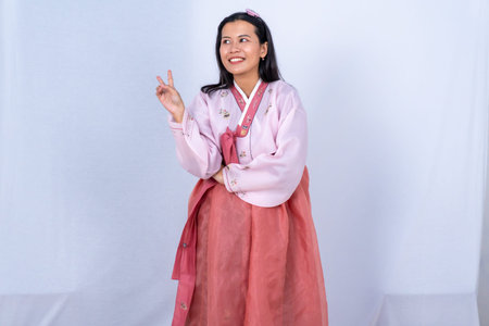 A woman in pink and red Korean Hanbok makes a peace sign with a smile against a white backgroundの写真素材